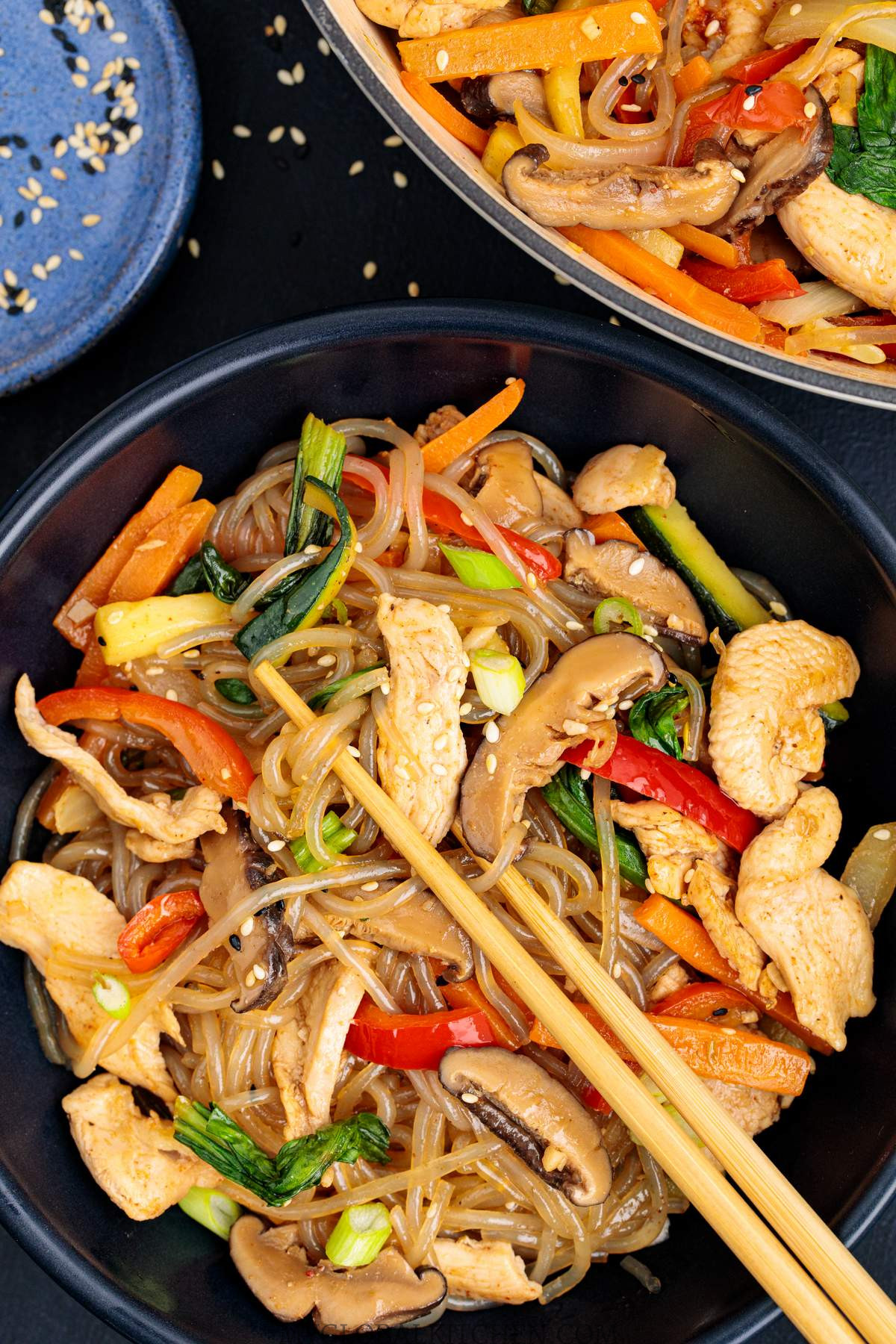 Closeup of Chicken Japchae stir-fry in black bowls with sesame seeds on the side