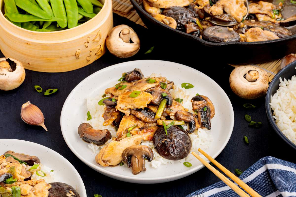 Rice and chinese chicken mushroom stir fry on a white plate with a skillet and vegetables in the backgroun