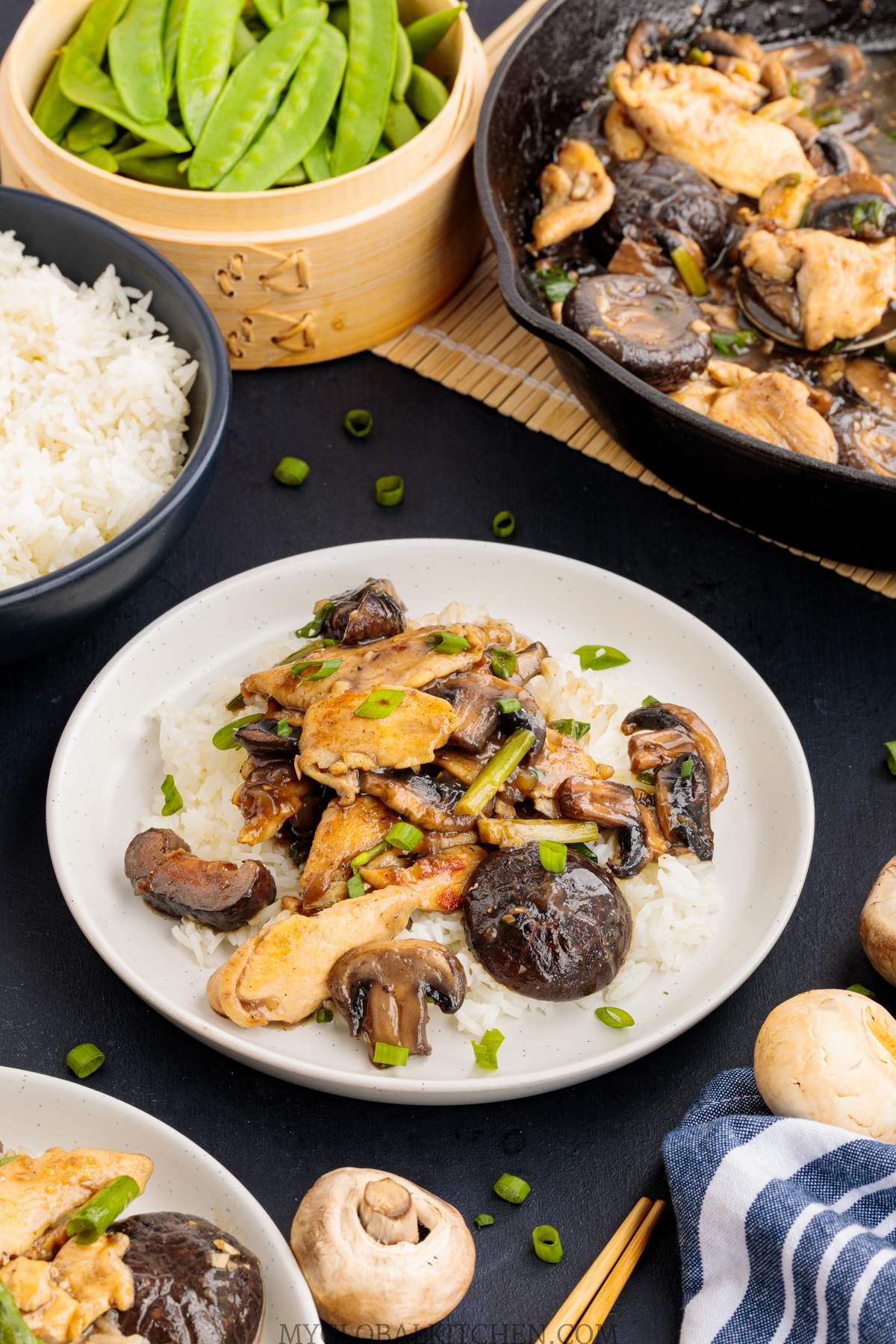 A white plate with rice and chinese chicken mushroom stir fry with a skillet and more rice in the backgroun