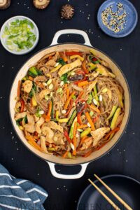 Overhead shot of a pot full of Korean Chicken Japchae or Glass Noodle Stir-Fry on a dark table with bowls of green onions and sesame seeds on the side