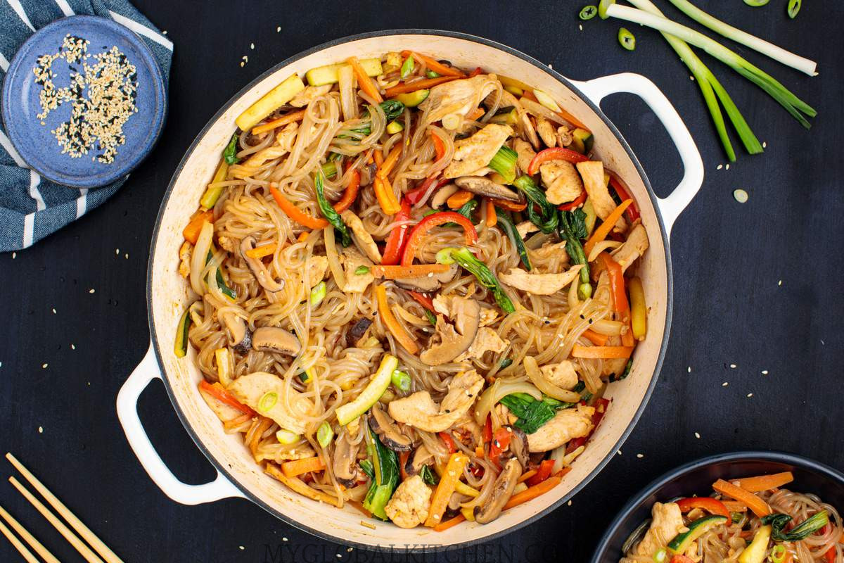 Korean Chicken Japchae or Glass Noodle Stir-Fry in a large pot on a dark table with bowls of green onions and sesame seeds on the side