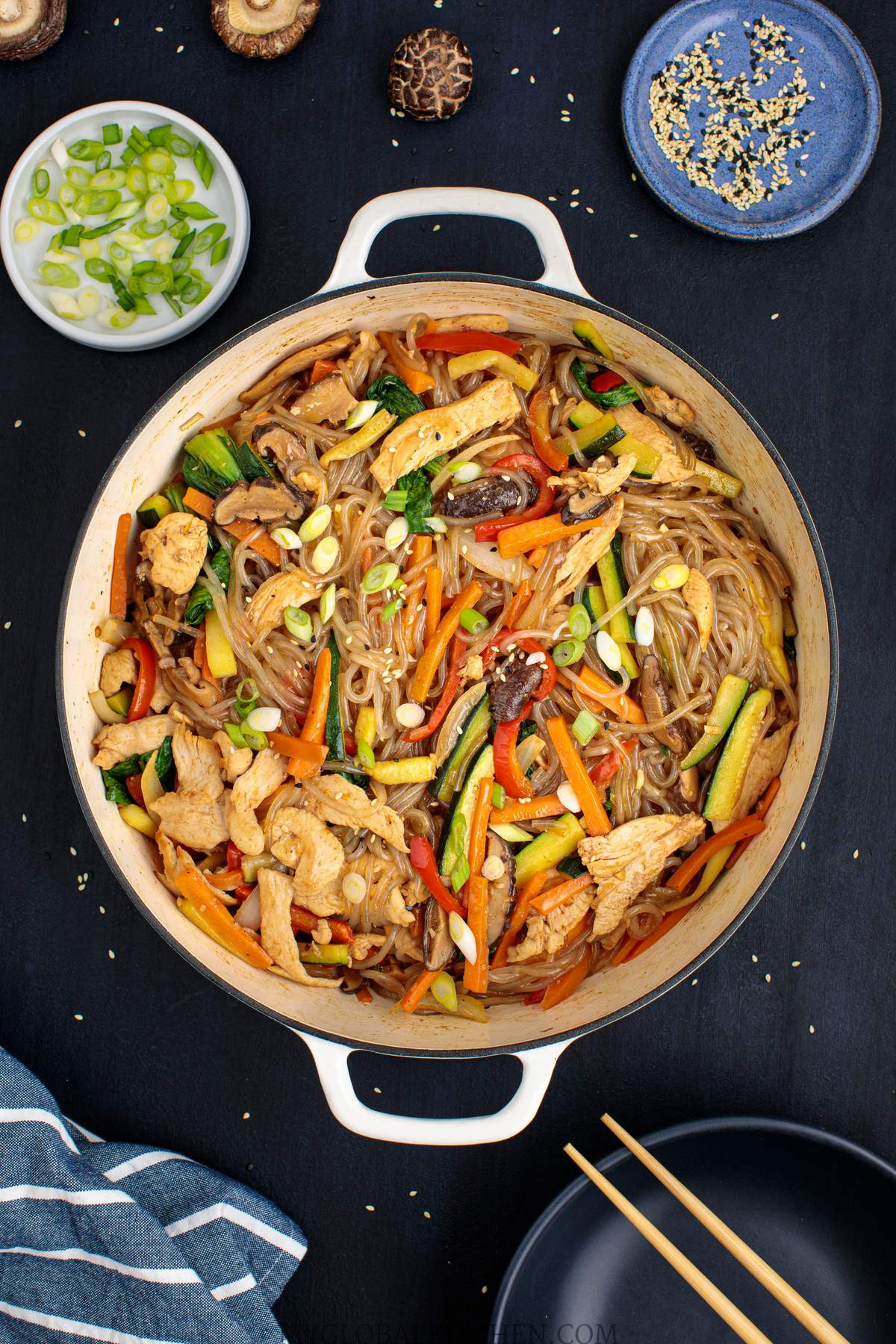 Overhead shot of a pot full of Korean Chicken Japchae or Glass Noodle Stir-Fry on a dark table with bowls of green onions and sesame seeds on the side