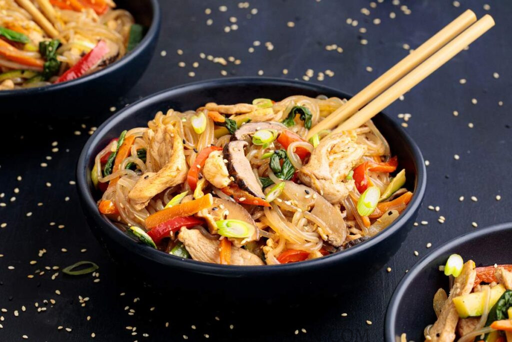 A black bowl filled with  Korean Japchae Glass Noodle Stir-Fry with Chicken, on top the bowl are a set of chopsticks