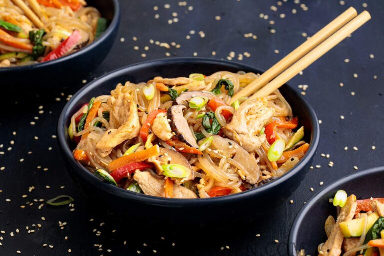 A black bowl filled with Korean Japchae Glass Noodle Stir-Fry with Chicken, on top the bowl are a set of chopsticks